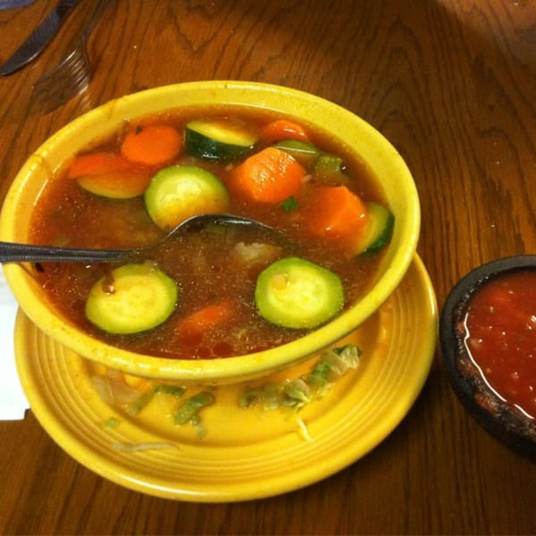 A yellow bowl of Mexican vegetable soup with carrots and zucchini, next to a small bowl of salsa.