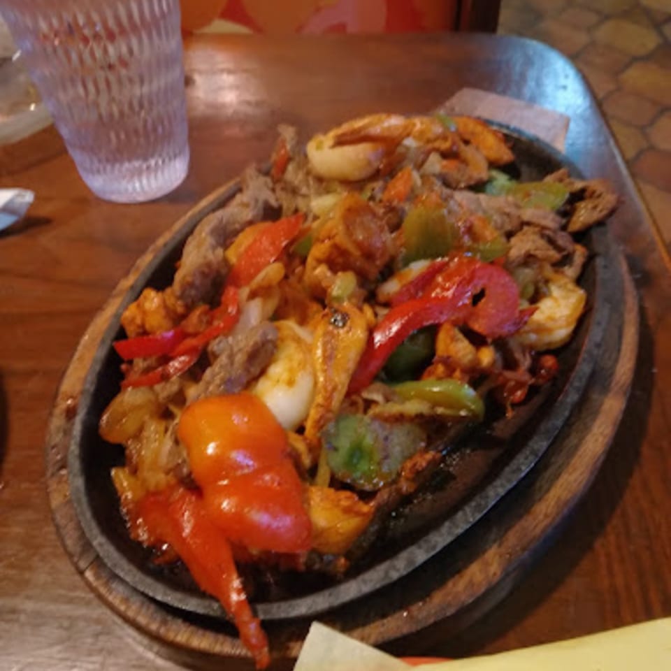 Sizzling fajitas with steak, shrimp, red and green bell peppers, and onions on a wooden platter.