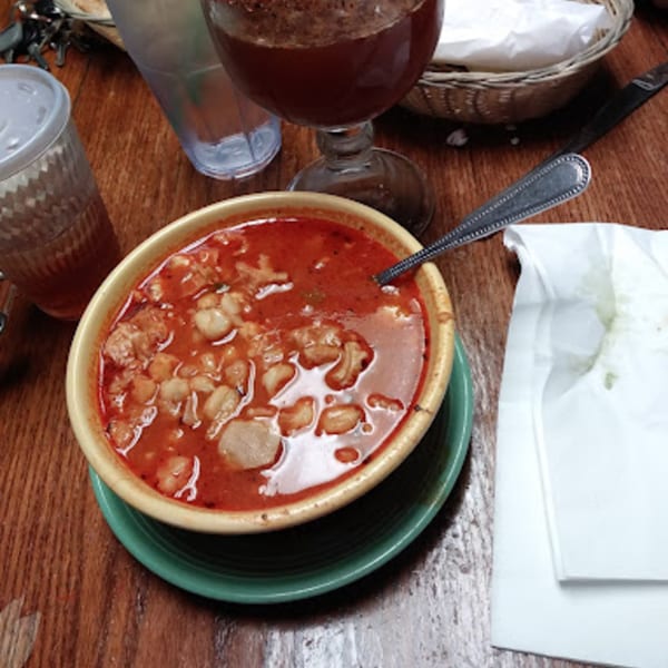 A bowl of red pozole soup with hominy and meat, a drink, and a napkin on a wooden table.