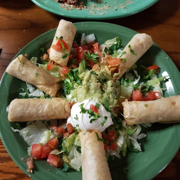 Crispy taquitos on a green plate, surrounded by lettuce, diced tomatoes, guacamole, and sour cream.